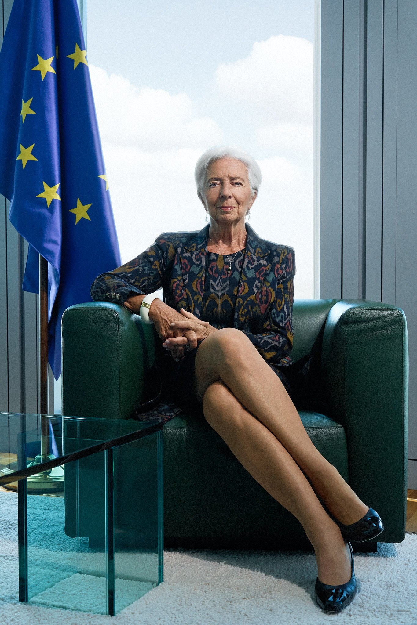 Eine Frau mit kurzen weißen Haaren sitzt auf einem dunkelgrünen Sessel mit gekreuzten Beinen in der Nähe einer EU-Flagge in einer modernen Büroumgebung. Sebastian Schueler – Business Fotograf Frankfurt am Main Christine Lagarde EZB Business Portrait Fotograf Frankfurt Sebastian Schueler